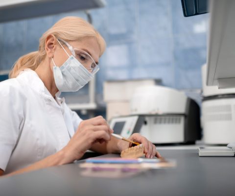 Concentrated dental technician wearing glasses and surgical mask making artificial teeth sitting in her laboratory